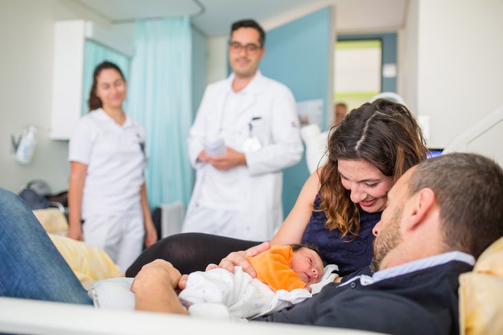 Frisch gebackene Eltern mit Baby, im Hintergrund zwei Ärzte